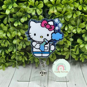 May include: A badge reel featuring a white Hello Kitty design with a pink bow, holding a blue beaker and cloud. The badge has a glittery blue border and a clear plastic backing. The text "Crafty Promises" is on the bottom.