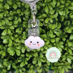 Puede incluir: Un dije de carrete para placa en forma de nube blanca con una cara sonriente y mejillas rosadas. El dije está unido a un carrete para placa de plástico transparente con un clip de metal plateado. El fondo es una densa disposición de hojas verdes.