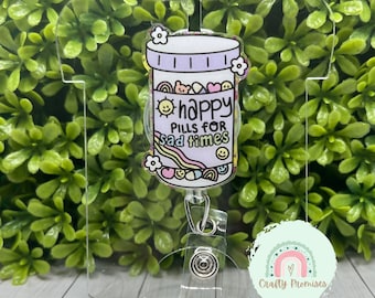 May include: A clear plastic badge holder with a colorful cartoon pill bottle charm. The pill bottle has the text "happy pills for sad times" and is decorated with rainbows, hearts, and smiley faces.