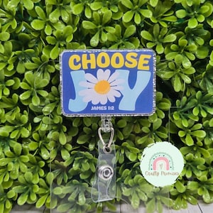 Puede incluir: Una placa rectangular con las palabras "CHOOSE JOY" en amarillo y azul, con una ilustración de margarita. El texto "JAMES 1:2" está debajo de la margarita. La placa está en un soporte de plástico transparente con un carrete retráctil. El fondo es una planta verde frondosa.