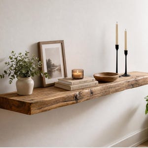 Könnte beinhalten: Rustikales, schwebendes Holzregal mit natürlicher Kante, dekoriert mit Wohnaccessoires. Enthält einen gerahmten Landschaftsdruck, Bücher, eine Kerze, eine Holzschale, schwarze Kerzenhalter mit Kerzen und eine Vase mit Grün.