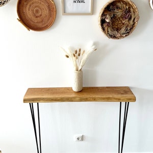 May include: A wooden console table with black hairpin legs. The table is made of natural wood with a live edge. The table is decorated with a vase of dried pampas grass.
