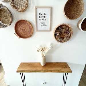 May include: A white wall with a framed print that reads "Heute nichts erlebt. Auch sch&ouml;n."  The print is surrounded by woven baskets of various shapes and sizes. A wooden console table with black hairpin legs sits in front of the wall. A vase with dried pampas grass sits on the table.