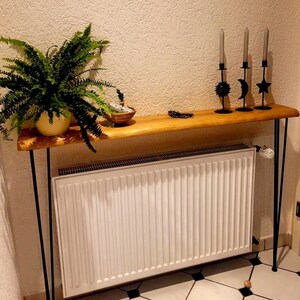 May include: A wooden console table with black hairpin legs. The table is placed over a white radiator and has a green plant in a pot on the left side. There are three black candle holders with a sun, moon, and star design on the table.
