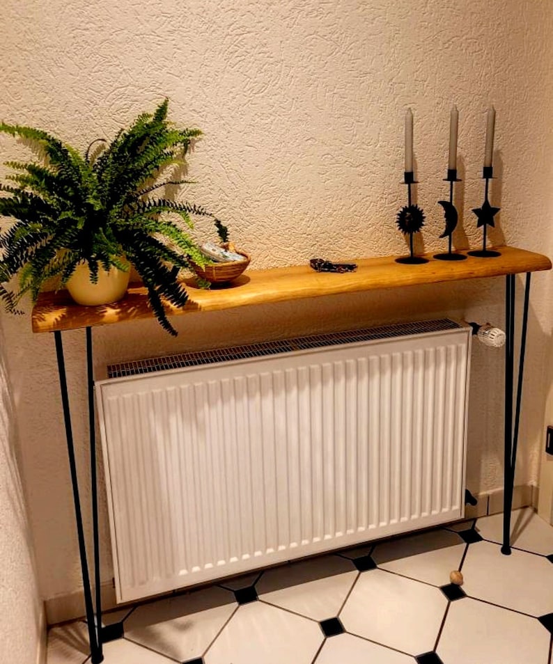 May include: A wooden console table with black hairpin legs. The table is placed over a white radiator and has a green plant in a pot on the left side. There are three black candle holders with a sun, moon, and star design on the table.