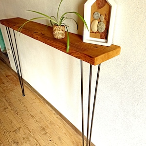 Può includere: Un tavolo console in legno con una finitura rustica e gambe a forcina. Il tavolo è decorato con una pianta in vaso e un quadro incorniciato di pietre e foglie.