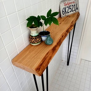 May include: A wooden console table with a live edge top and black hairpin legs. The table is decorated with a small potted plant, a ceramic vase, and a California license plate that reads "6K25214".
