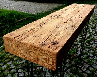 Puede incluir: Una mesa de madera rústica con un acabado de madera natural y un aspecto desgastado. La mesa tiene una tapa rectangular y está sostenida por cuatro patas de horquilla negras. La mesa está sentada sobre una cama de grava y hierba.