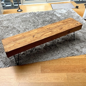Könnte beinhalten: Eine lange Holzbank mit naturbelassenem Holzfinish und schwarzen Haarnadelbeinen. Die Bank steht auf einem grauen Shaggy-Teppich.