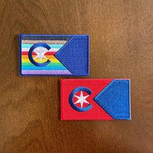 May include: Two rectangular embroidered patches. The top patch features horizontal stripes in black, brown, white, red, orange, yellow, green, blue, and violet, with a blue triangle on the right and a blue letter C enclosing a white star. The bottom patch is solid red with a blue triangle and a blue letter C enclosing a white star.