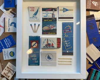 May include: A white shadow box frame containing a collection of vintage matchbooks and loose matches. The display features various nautical-themed matchbook covers with logos and illustrations of boats, lighthouses, and yacht clubs arranged on a white background.