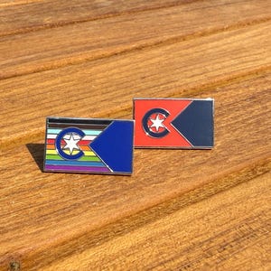 May include: Two rectangular enamel pins featuring a stylized flag design. One pin displays horizontal rainbow stripes with a blue triangle and a central circle containing a white star. The second pin features a solid red field with a blue triangle.