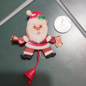 May include: A plastic Santa Claus ornament with a red hat, white beard, and red suit, holding a small bell and a sign that reads Merry Christmas. A red string with a bell hangs from the bottom, and a US quarter is shown for scale.