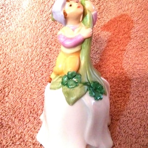 May include: A ceramic figurine of a child wearing a lavender hat and yellow pants, embracing a green stalk. The figurine is perched atop a white, petal-shaped base, with green shamrock accents.