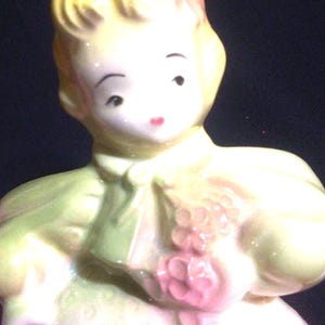 May include: A porcelain figurine of a girl with blonde hair wearing a green dress and holding a bouquet of pink flowers.