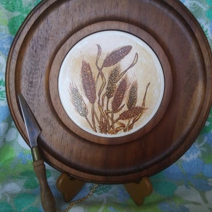 May include: A wooden cheese board with a round, ceramic tile insert featuring a brown and white wheat design. The board has a metal chain attached to a small knife.
