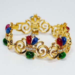 May include: A gold bracelet with a delicate, ornate design. The bracelet features a repeating pattern of crowns with red, blue, and green gemstones.