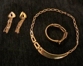May include: A set of gold-toned jewelry featuring a necklace, bracelet, and earrings. The necklace has a curved design with a chain link pattern. The bracelet is a simple chain link design. The earrings are small and have a dangling chain.
