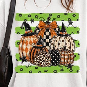 May include: A white long-sleeve sweatshirt featuring a graphic print of several pumpkins with various patterns including leopard print, polka dots, and plaid. Some pumpkins wear witch hats, and the design is set against a lime green striped background.