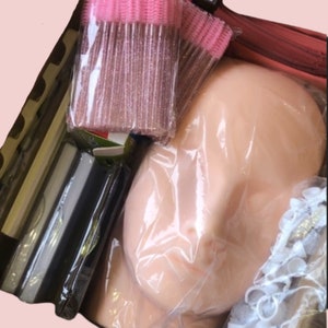 May include: A close-up of a makeup practice head with a pink and white plastic lash applicator, a clear plastic bag, and a white plastic container with a white and pink label.
