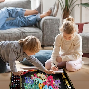 May include: A black and gold Football Stadium map with colorful stadium icons. Two children are interacting with the map, one using a pen and the other with a toy car. The map is on a wooden floor.