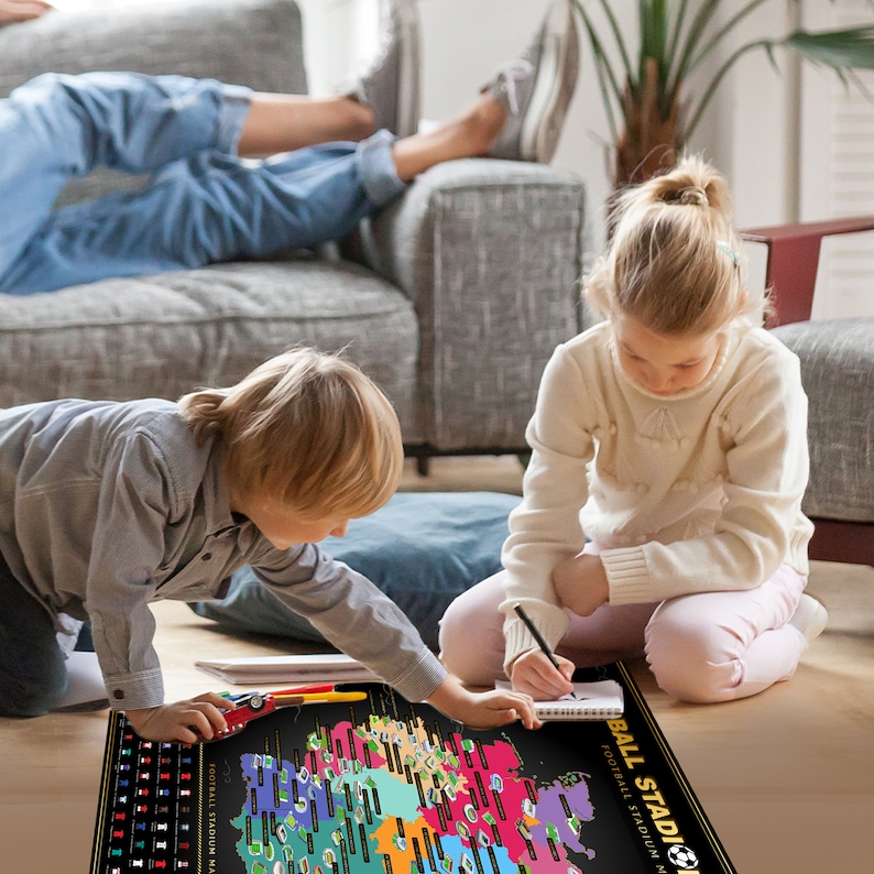 May include: A black and gold Football Stadium map with colorful stadium icons. Two children are interacting with the map, one using a pen and the other with a toy car. The map is on a wooden floor.