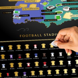 May include: A black and gold Football Stadiums scratch-off map with team jerseys and stadium illustrations. The map includes the text "FOOTBALL STADIUM". A hand is peeling off a sticker from the map.