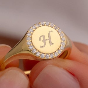 Könnte beinhalten: Ein goldener Siegelring mit einer runden Fläche, auf der ein einzelnes Initial "H" in der Mitte graviert ist. Die Fläche ist von einem Heiligenschein aus kleinen, klaren Zirkonia-Steinen umgeben.