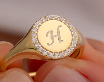 May include: A gold signet ring with a round face featuring a single initial "H" engraved in the center. The face is surrounded by a halo of small clear cubic zirconia stones.