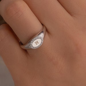 May include: A silver oval signet ring with a single initial engraved in the centre and a row of small diamonds around the edge.