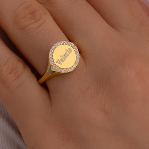 May include: A gold signet ring featuring a circular face engraved with the name Valerie. The center of the ring is surrounded by a border of small, clear, sparkling stones set into the gold band, creating a polished and reflective appearance.