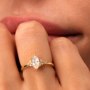 May include: A gold ring featuring a central marquise-cut clear gemstone flanked by two smaller round-cut clear stones on each side. The band is thin and polished, showcasing a classic design with a prong setting for the stones.