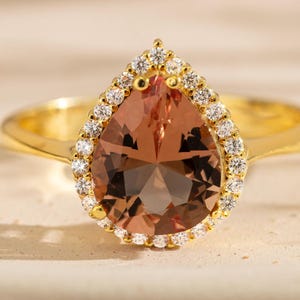 May include: A gold ring featuring a large, pear-shaped, peach-colored gemstone at the center. The central stone is surrounded by a halo of small, clear, round-cut stones set in a gold band that tapers toward the back of the ring.