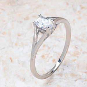 May include: A silver engagement ring with a large, clear, oval-cut gemstone. The ring has a delicate band and a split shank design, with the gemstone held in place by four prongs. The ring is set against a neutral background.