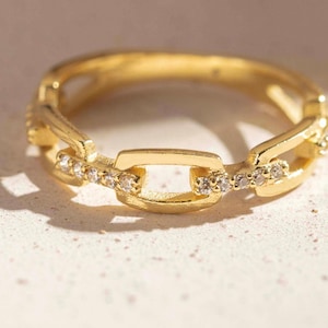 Könnte beinhalten: Ein goldfarbener Ring mit einem Gliederkettendesign. Das Band besteht aus abwechselnd glatten Metallgliedern und Gliedern, die mit kleinen, klaren, funkelnden Steinen besetzt sind. Der Ring hat eine polierte Oberfläche und eine runde Form.