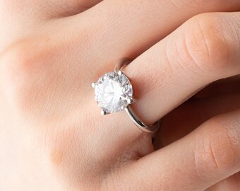 May include: A silver ring with a large, clear, round diamond set in a prong setting. The ring is on a person's finger, and the person's nails are painted a dark green.