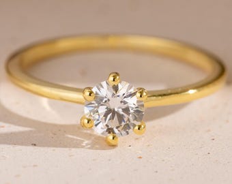 May include: A gold ring featuring a single round, clear, faceted gemstone held in place by a six-prong setting. The band is smooth and polished, showcasing a bright yellow metallic finish that contrasts with the brilliant white stone at the center.