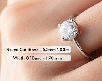May include: A silver engagement ring with a round, clear diamond. The ring has a band that is 1.70 mm wide. The diamond is 6.5 mm in diameter and 1.00 carats.