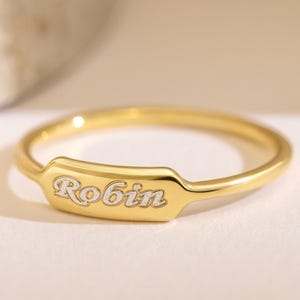 Peut inclure: Une bague dorée dotée d'un anneau fin et poli ainsi que d'une plaque rectangulaire centrale. La plaque est gravée du prénom Robin dans une police cursive, avec des lettres présentant une finition métallique plus claire et contrastée.