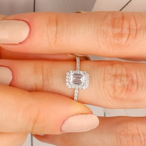 May include: A close-up of a hand wearing a silver ring with a square-shaped diamond surrounded by smaller diamonds.