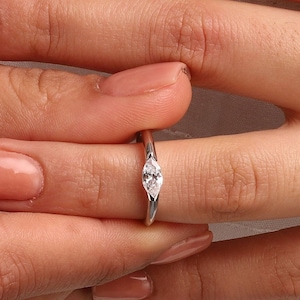 Può includere: Un anello d'argento con un solo diamante rotondo trasparente incastonato a griffe. L'anello è su un dito.