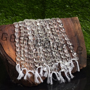 Puede incluir: Hilos de cuentas de piedras preciosas claras y facetadas con borlas blancas. Las cuentas son redondas y tienen una superficie pulida y reflectante. Las piedras preciosas se exhiben sobre una superficie de madera.