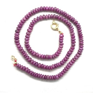 May include: A necklace composed of small, rounded, purplish-pink beads. The beads are strung together, forming a circular shape. The necklace features a gold-colored clasp. The jewelry is displayed against a white background.