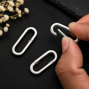 May include: Three silver-toned metal oval carabiner clips with a spring-loaded gate mechanism. Two of the carabiners are lying flat, while one is being held to demonstrate the opening function of the hinged gate on the side of the oval frame.