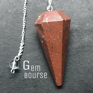 May include: A faceted, cone-shaped red jasper pendulum suspended from a silver-coloured metal chain. The stone features dark brown veining and a smooth, polished surface. A small, clear bead is attached to the chain. The text Gem bourse is visible.