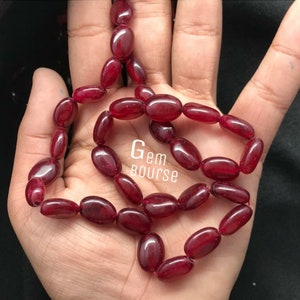 May include: A strand of deep red oval-shaped gemstone beads. The beads are strung on a clear thread and are approximately 1 cm in length. The beads are displayed on a hand.