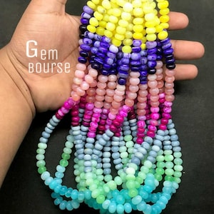 May include: Strands of colorful gemstone beads in a variety of colors, including yellow, purple, pink, and blue. The beads are small and round, and the image includes the text "Gem Bourse".