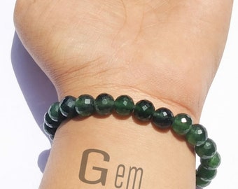 Pode incluir: Uma pulseira composta por contas de pedras preciosas redondas, facetadas e de cor verde escura. As contas estão enfiadas juntas num único fio. O texto "Gem BOURSE" é visível na imagem, posicionado abaixo da pulseira na superfície da pele.