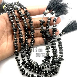 May include: A strand of black gemstone beads with white spacers. The beads are irregular in shape and have a rough, natural texture. The strand is finished with black tassels.