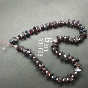 Könnte beinhalten: Eine Halskette aus grob geschnittenen, dunkelroten Granatsteinen, die mit kleinen weißen Perlen durchsetzt sind. Die Halskette ist auf einer dünnen, rötlich-braunen Schnur aufgefädelt. Der Hintergrund ist eine dunkelgraue Oberfläche.